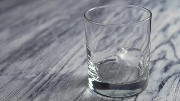 Zmrazené kostky ledu padají do prázdné sklenice na whisky na staré dřevěné šedé barové lince - Záběry, video