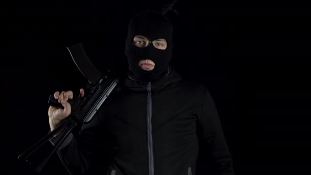 Ein Mann mit Sturmhaube steht mit einem Sturmgewehr vom Typ AK-47. Der Räuber steht mit einem Gewehr auf der Schulter und zielt dann in die Kamera. Auf schwarzem Hintergrund. - Filmmaterial, Video