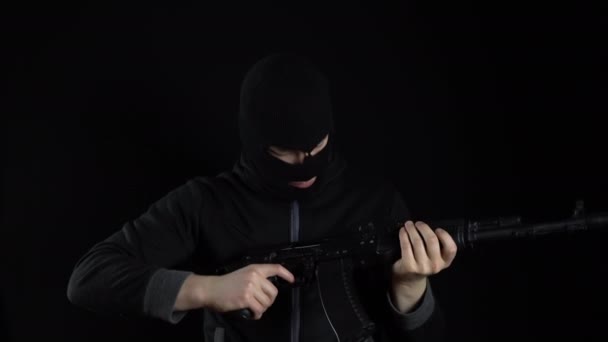 Een man met een bivakmuts staat met een AK-47 aanvalsgeweer. De bandiet laadt de machine op en staat. Op een zwarte achtergrond. - Video