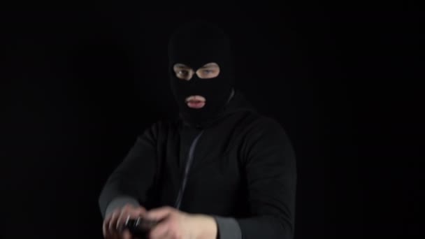 Um homem com uma máscara de balaclava está com uma arma. Um bandido aponta a arma para a câmara. Em um fundo preto. - Filmagem, Vídeo
