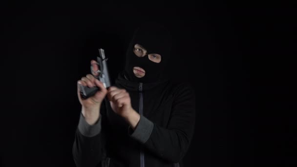 Een man met een bivakmuts staat met een pistool. De bandiet laadt het pistool opnieuw op en staat op een zwarte achtergrond. - Video
