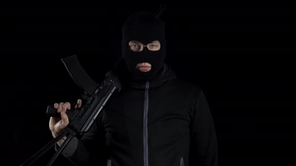 Kar maskeli bir adam AK-47 saldırı tüfeğiyle ayakta duruyor. Siyah bir arka planda, omzunda silahla bir haydut duruyor.. - Video, Çekim
