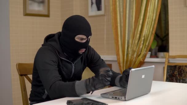 Un ladrón está tratando de hackear un portátil. Un matón enmascarado está sentado en una casa tratando de entrar en una computadora portátil y está amenazando con un arma. Robo de datos desde un ordenador. - Imágenes, Vídeo