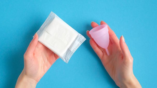 Female hands holding menstrual cup and sanitary napkin over blue background. Different types of menstrual hygiene products comparison concept - Φωτογραφία, εικόνα