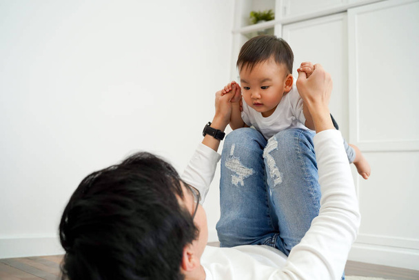 Joyful Asian boy playing airplane with parenting dad in the living room at home. Father is carrying innocent son up on his legs ready to fly. Fatherhood in Asia concept - Photo, Image