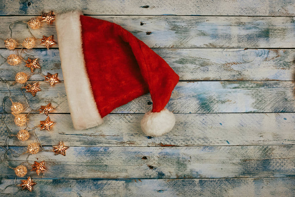 Рождественский фон с шапкой Санты и рождественскими огнями на деревенском винтажном деревянном фоне. Гарланд
.  - Фото, изображение