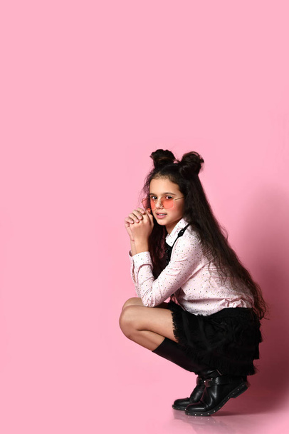 Teenage kid with long hair, in sunglasses, white blouse, black skirt and boots. She posing squatting on pink background. Close up - Photo, image