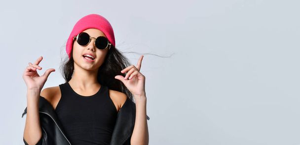 Έφηβη γυναίκα με μαύρο δερμάτινο μπουφάν ροζ καπέλο και γυαλιά ηλίου. Κάνει χειρονομίες ενώ ποζάρει απομονωμένη πάνω στο λευκό. Κλείσε. - Φωτογραφία, εικόνα