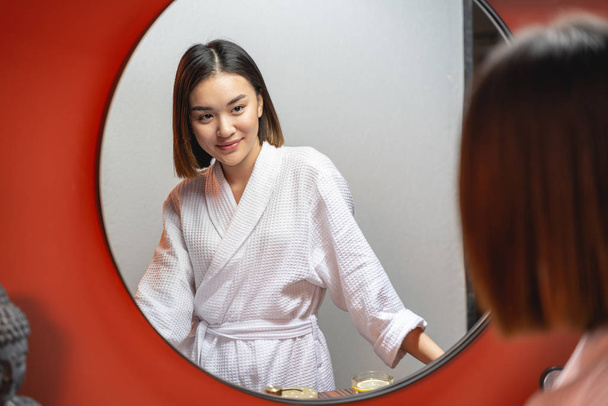 Молода азійська дівчина дивиться на дзеркало у приміщенні. - Фото, зображення