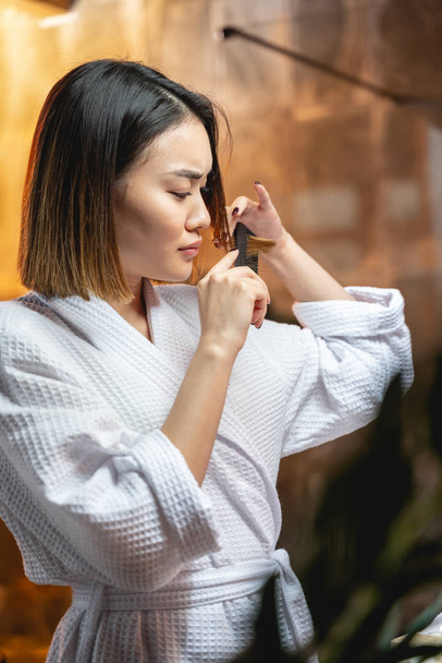 Sad Asian lady in white bathrobe brushing her hair - 写真・画像