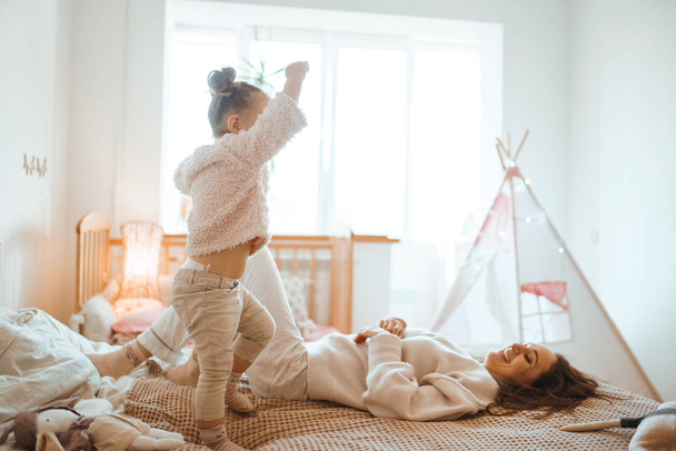 Happy young woman and her little cute daughter are having fun in bed while being at home together. Mother playing and jumping with her baby in the bedroom. Happy Mother's Day. Happy loving family.  - 写真・画像