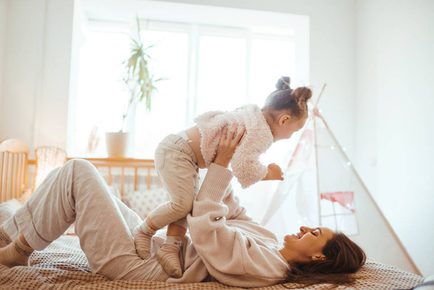 Happy young woman and her little cute daughter are having fun in bed while being at home together. Mother playing and jumping with her baby in the bedroom. Happy Mother's Day. Happy loving family.  - Photo, image