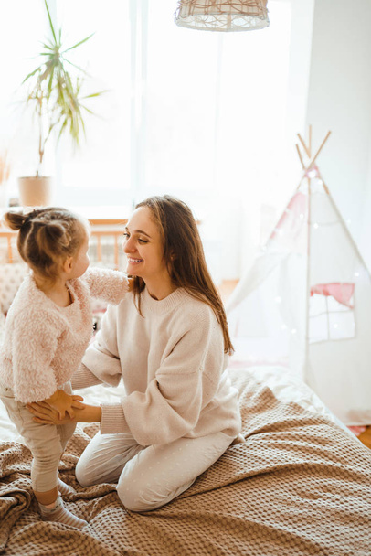 Happy young woman and her little cute daughter are having fun in bed while being at home together. Mother playing and jumping with her baby in the bedroom. Happy Mother's Day. Happy loving family.  - Фото, зображення