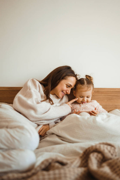 Cheerful mother and her daughter little daughter watching cartoons on phone lying In bed at home. Happy loving family. Modern wireless tech usage free time concept. Bedtime Fun. - Foto, Imagen