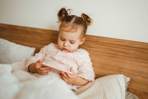 Happy little girl watching cartoons on phone lying In bed at home. Happy loving family. Modern wireless tech usage free time concept. Bedtime Fun. - Foto, Imagen