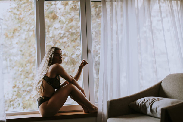 Молодая женщина в нижнем белье сидит и смотрит в окно своей красивой квартиры
 - Фото, изображение
