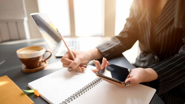 Widok boczny bizneswoman pisząc na notebooku podczas korzystania ze smartfona z laptopem i filiżanką kawy w komfortowym miejscu pracy - Zdjęcie, obraz