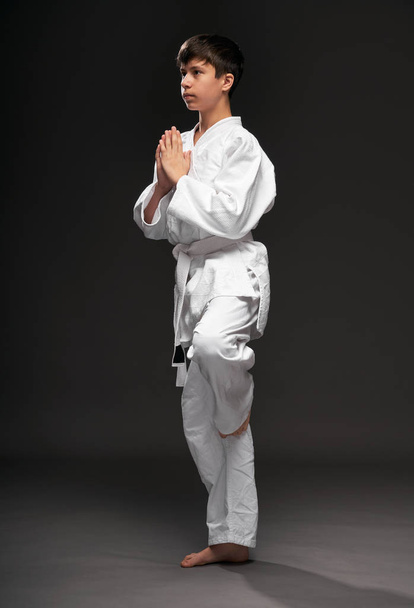 um adolescente vestido com roupas de artes marciais posa em um fundo cinza escuro, um conceito esportivo
 - Foto, Imagem