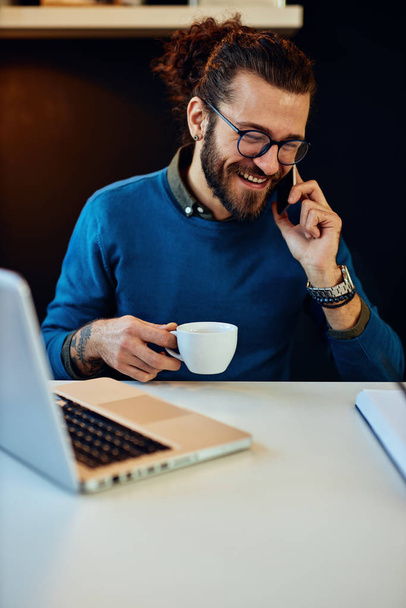 Jeune blogueur barbu caucasien souriant assis dans son bureau, parlant au téléphone, tenant une tasse de café et prenant une pause du travail
. - Photo, image