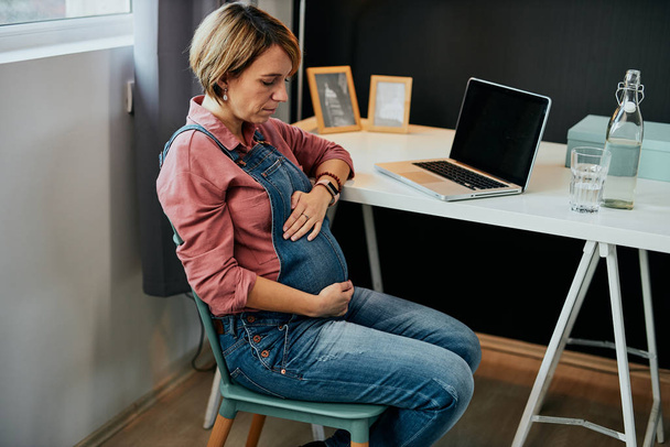 Fière future mère assise dans son bureau à la maison et touchant le ventre
. - Photo, image