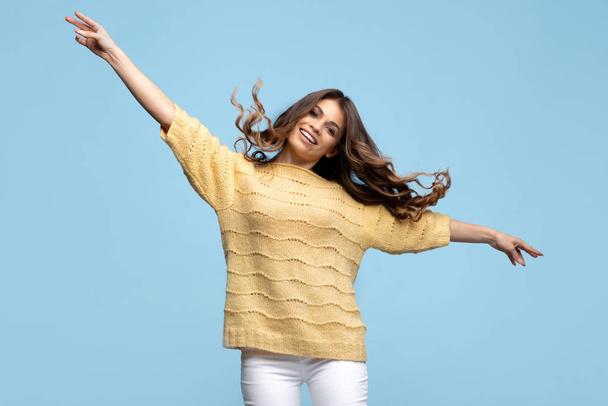 Happy young woman in yellow sweater dancing with hands up over blue background. - Φωτογραφία, εικόνα