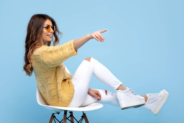Happy young woman in yellow sweater pointing over blue background. Pretty girl in yellow glasses. - Foto, Imagen
