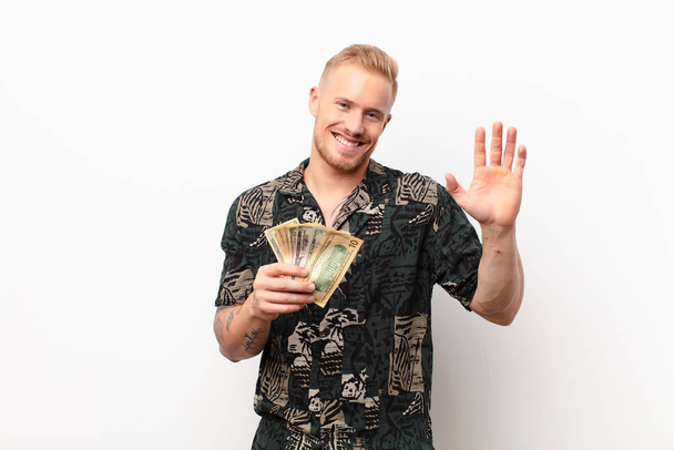 young blonde man smiling happily and cheerfully, waving hand, welcoming and greeting you, or saying goodbye against white wall with banknotes - Fotoğraf, Görsel