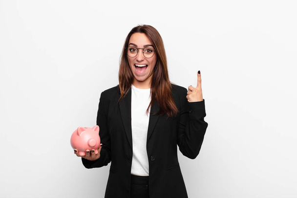 young businesswoman feeling like a happy and excited genius after realizing an idea, cheerfully raising finger, eureka! with a piggy bank - Foto, Imagen