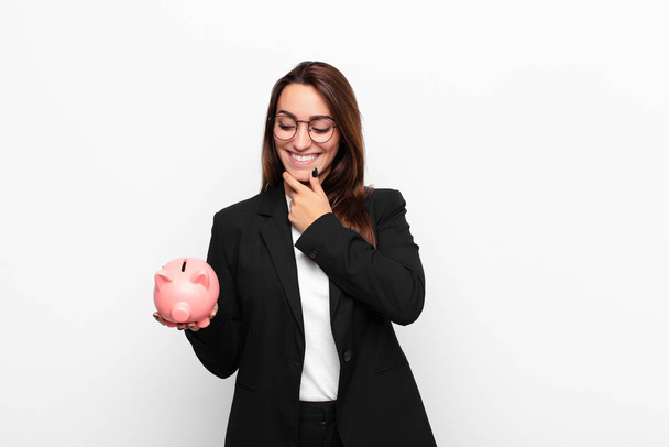 young businesswoman smiling with a happy, confident expression with hand on chin, wondering and looking to the side with a piggy bank - Photo, Image