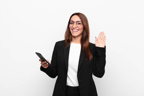 young businesswoman smiling happily and cheerfully, waving hand, welcoming and greeting you, or saying goodbye and holding a mobile telephone - Fotografie, Obrázek