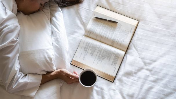 コーヒーを飲む時間だ。アジアの怠惰な女性が横たわって、柔らかい朝の光で午前中に本を読んだ後、ベッドの上にコーヒーのカップを保持する。朝のライフスタイルのコンセプト. - 写真・画像