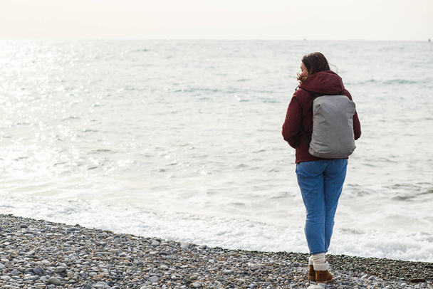 Een vrouw in een jas met een rugzak kijkt naar de zee golven uitzicht van achteren - Foto, afbeelding