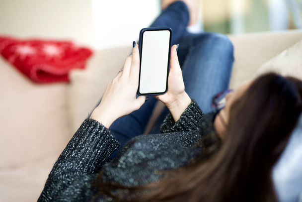Rear view shot of young woman holding mobile phone in her hand while relaxing on the sofa. - Foto, afbeelding