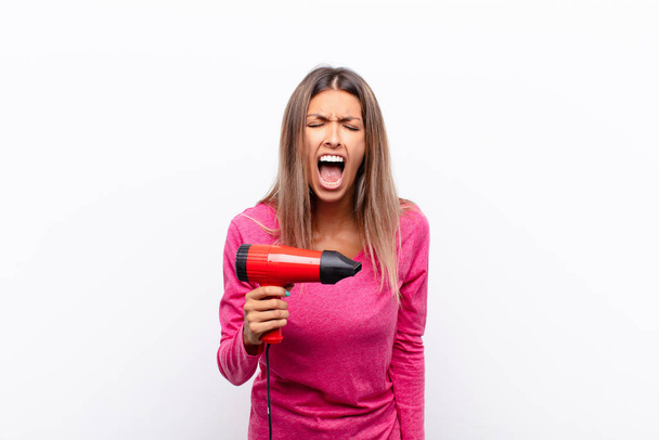 young pretty woman shouting aggressively, looking very angry, frustrated, outraged or annoyed, screaming no with a hair dryer. - Fotoğraf, Görsel