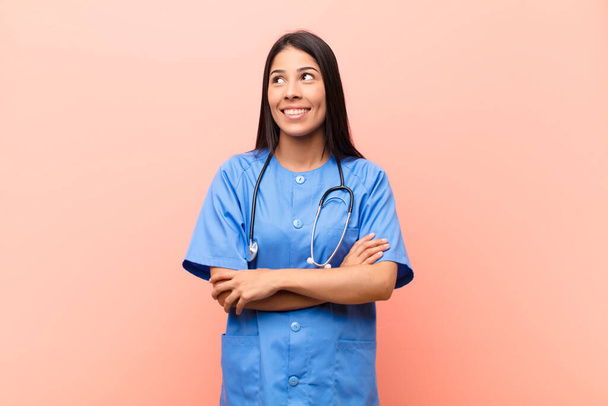 young latin nurse feeling happy, proud and hopeful, wondering or thinking, looking up to copy space with crossed arms against pink wall - Foto, Imagen