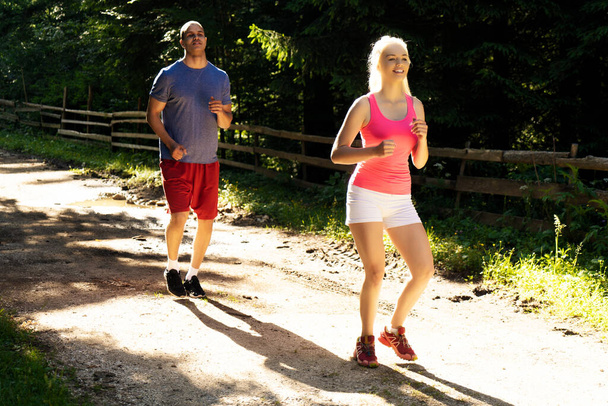 Running fitness couple of runners doing sport on road outdoor. Active living man and woman jogging training cardio in summer outdoors nature. - Photo, Image