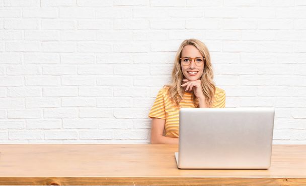 young blonde woman looking happy and smiling with hand on chin, wondering or asking a question, comparing options using a laptop - Foto, Bild