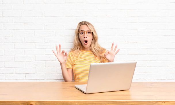young blonde woman feeling shocked, amazed and surprised, showing approval making okay sign with both hands using a laptop - Фото, изображение