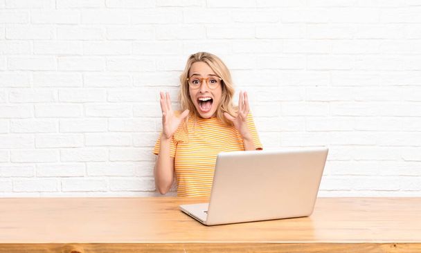 young blonde woman feeling shocked and excited, laughing, amazed and happy because of an unexpected surprise using a laptop - Fotó, kép