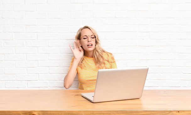 young blonde woman looking serious and curious, listening, trying to hear a secret conversation or gossip, eavesdropping using a laptop - Photo, image