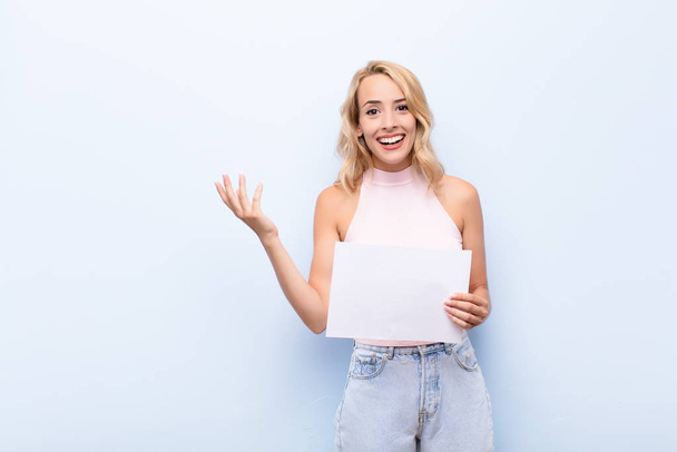 young blonde woman feeling happy, surprised and cheerful, smiling with positive attitude, realizing a solution or idea holding a sheet of paper - Fotó, kép