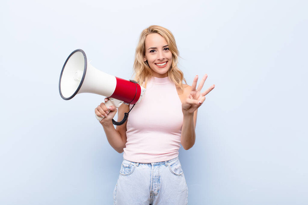 young blonde woman smiling and looking friendly, showing number three or third with hand forward, counting down with a megaphone - Foto, imagen