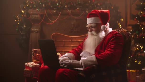 An elderly man in a Santa Claus costume sits in a beam of light at home against the background of a fireplace and a Christmas tree on Christmas eve and types on a laptop while sitting on the Internet. - Filmagem, Vídeo