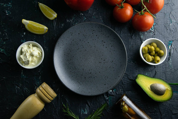 Grijze plaat met duidelijke ruimte op een zwarte achtergrond, rond de tomaat, saus, olijven, citroen, olijfolie met specerijen. - Foto, afbeelding