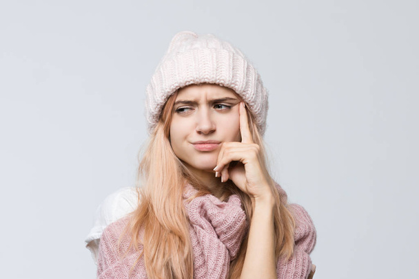 Indoor portrait of cute girl in a pink sweater and white hat frowning face, presses hand to cheek, looks aside with pensive expression/ doubts, trying to remember something important, emotions concept - Zdjęcie, obraz