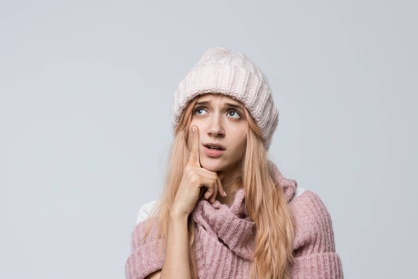 Studio photo of attractive teen in a pink sweater and white hat deep in thought, lost in thought, looks aside, presses lips together/ pensive woman, looking helpless and disoriented, emotions concept. - Photo, Image