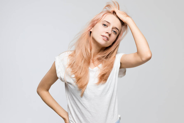 Studio portrait of cute young blonde woman in white t-shirt frowning her face in displeasure, straightens hair with hand/closed posture, angry, strict and skeptical expression, negative emotion. - 写真・画像