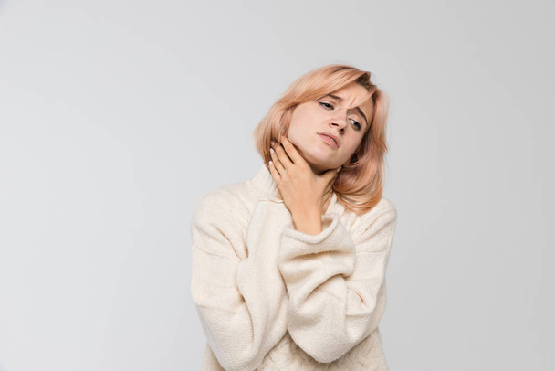 Studio portrait of young unhealthy blonde woman in warm sweater holding her throat/suffers from cold and flu, severe sore throat, feeling sick, chest pain, medicine and healthcare concept. - Photo, Image