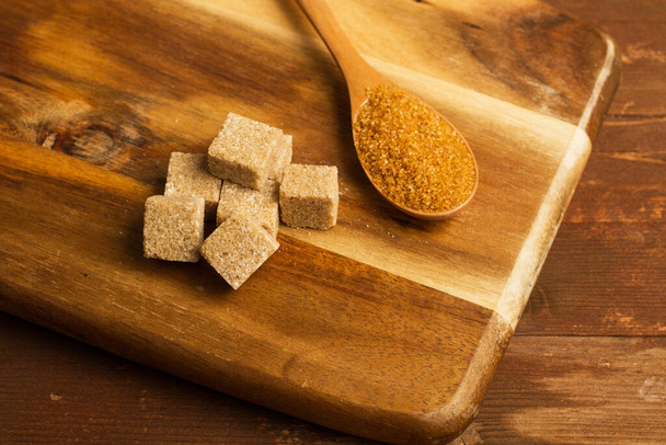 Cubos de azúcar moreno en una tabla de cocina de madera y una cuchara de madera con azúcar morena
 - Foto, Imagen