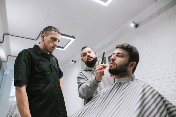 Two barbers cut a client with a beard at a men's hairdresser. Male teacher teaches a student to do hairstyles on a model. Workshop in Barbershop. Workshop on Men's Hairstyles Concept. - 写真・画像